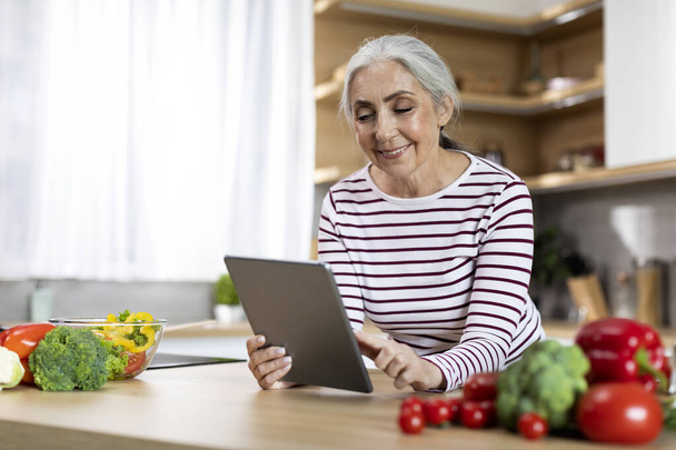 Portrait Of Happy Beautiful Senior Woman Using Digital Tablet While Cooking In Kitchen, Smiling Elderly Lady With Modern Gadget Checking Online Recipe Or Shopping In Internet, Copy Space - 写真・画像