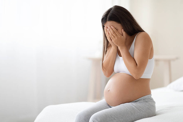 Pregnancy hormonal changes concept. Young long-haired pregnant woman in homewear with big tummy sitting alone on bed at home, covering her face, feeling sad and crying, free space - Zdjęcie, obraz