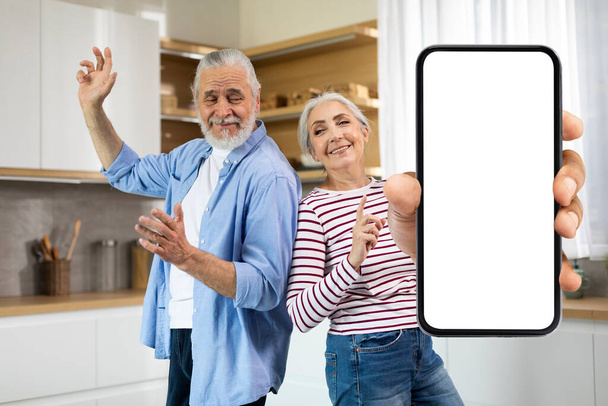 Joyful Senior Couple Dancing In Kitchen And Showing Big Blank Smartphone With White Screen At Camera, Positive Elderly Spouses Having Fun And Demonstrating Copy Space For App Design, Mockup - Zdjęcie, obraz
