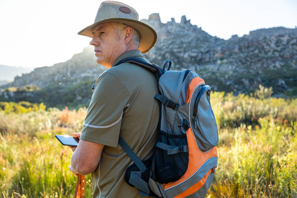 Зрілий чоловічий пішохід з рюкзаком у капелюсі та навігаційним напрямком над смартфоном, насолоджуючись відпусткою серед природи
. - Фото, зображення