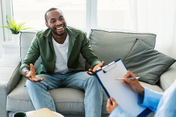 Effective psychotherapy. Happy black man talking to his psychologist, receiving professional help at mental health clinic. Male patient sharing therapy results with counselor - Fotografie, Obrázek