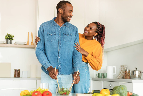 Cooking together. Happy loving black wife hugging husband while man cooking salad for dinner in kitchen at home. Spouses enjoying food preparation. Family nutrition and recipes - Foto, Imagen