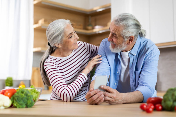 Сучасні технології. Щасливі старші подружжя використовують смартфон, розслабляючись на кухні разом, посміхаючись літнім чоловіком і дружиною тримаючи мобільний телефон і дивлячись один на одного, насолоджуючись новим додатком
 - Фото, зображення