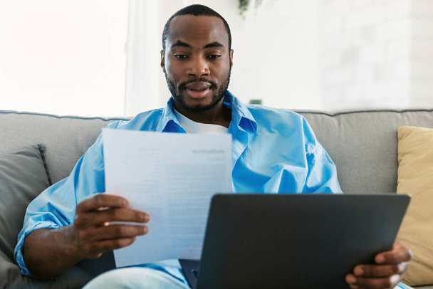 Remote career, freelance job concept. Young african american man using laptop computer and looking through business documents during online work at home - Foto, Bild