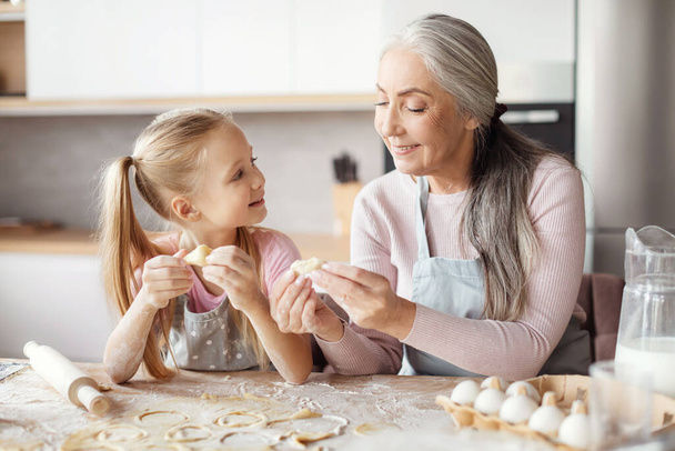 Smiling old grandmother in apron teaching little granddaughter making dough, mold cookies, prepare food in minimalist kitchen interior. Household chores together, cooking lesson at home, free space - Fotografie, Obrázek