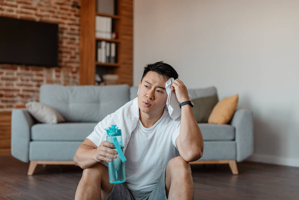 After training. Tired asian man with white towel on neck holding shaker with water and swiping sweat from forehead, resting after domestic training at home - Foto, Bild