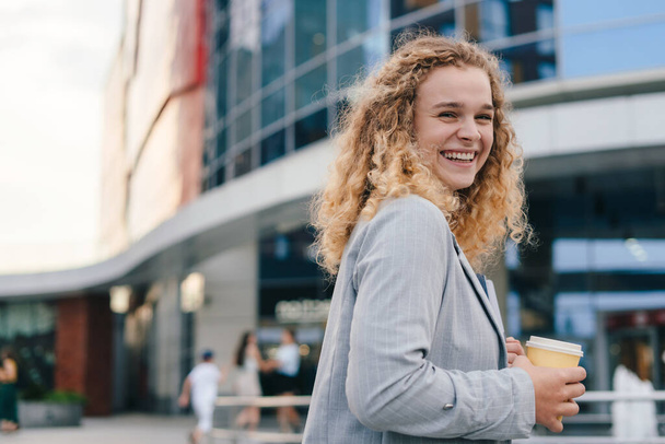 Щаслива молода жінка з кучерявим випивкою забирає каву, що йде по вулиці, дивлячись на камеру і посміхаючись. Концепція освіти. Люди міського способу життя
 - Фото, зображення