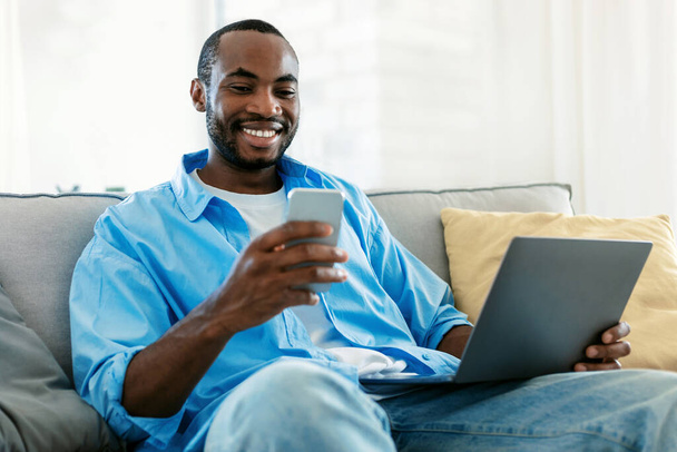 Business and telecommuting concept. Young black man using smartphone, sitting on sofa with laptop computer, reading text message, working at home office - Foto, Imagem