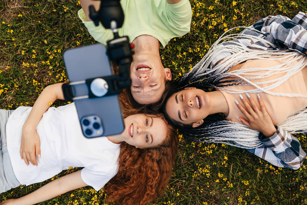 Happy three people lying down on green grass holding heads together looking up at the camera as posing for a selfie photo. New generation, gadgets easy usage in - Zdjęcie, obraz