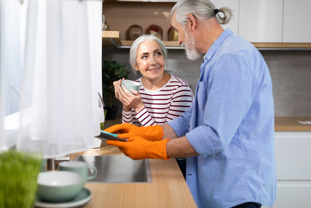 Caring Elderly Husband Helping Wife With Dishes In Kitchen, Smiling Senior Man Cleaning Plates While His Spouse Drinking Coffee, Happy Aged Spouses Sharing Domestic Chores, Free Space - Foto, afbeelding