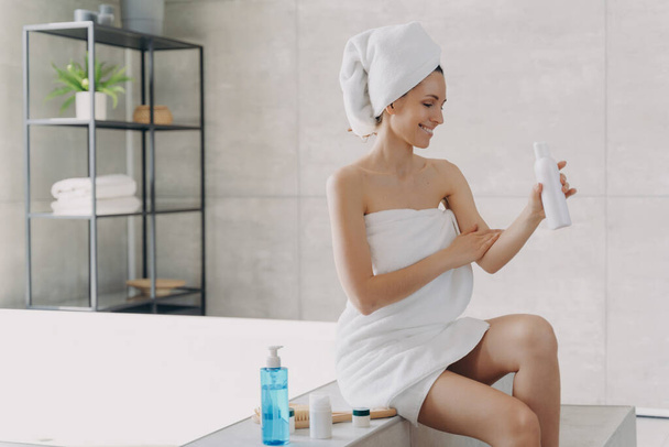 Smiling woman wrapped in towel applying organic body care lotion sitting on bathtub in modern bathroom. Female using sunscreen cream after shower. Self-care, skincare treatment, spa procedure - Foto, afbeelding