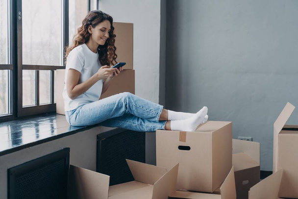 Modern girl homeowner tenant using smartphone app, choosing moving company, chatting with mover online, sitting on windowsill with cardboard boxes, waiting to move to a new house. Relocation concept. - Фото, изображение