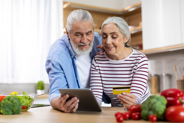 Smiling Senior Spouses Making Online Payments With Digital Tablet And Credit Card While Relaxing In Kitchen, Portrait Of Happy Elderly Couple Using Modern Gadget For Internet Shopping, Copy Space - Fotó, kép