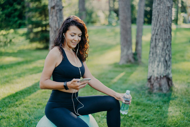 Angenehm entspannte Frau ruht sich nach Yoga-Übungen auf Fitnessball aus, hört Musik über modernes Handy und Kopfhörer, hält Flasche Frischwasser in der Hand, trägt Aktiv-Kleidung, posiert im Park - Foto, Bild