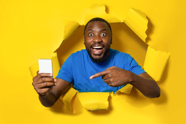 Shocked african american man holding cellphone and pointing on it, standing through yellow paper torn hole, looking at camera with open mouth - Foto, immagini