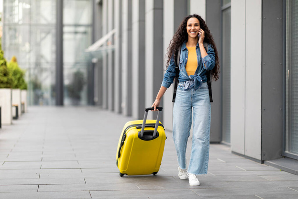Smiling young brunette curly woman in casual outfit traveller walking by airport, carrying yellow suitcase, having phone conversation, looking at copy space, panorama, full length - Foto, Bild