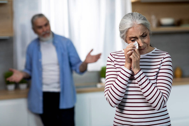 Depressed Senior Woman Crying In Kitchen During Argue With Husband, Upset Aged Female Wiping Tears With Napkin, Elderly Spouses Suffering Relationship Crisis And Misunderstanding, Selective Focus - Фото, изображение