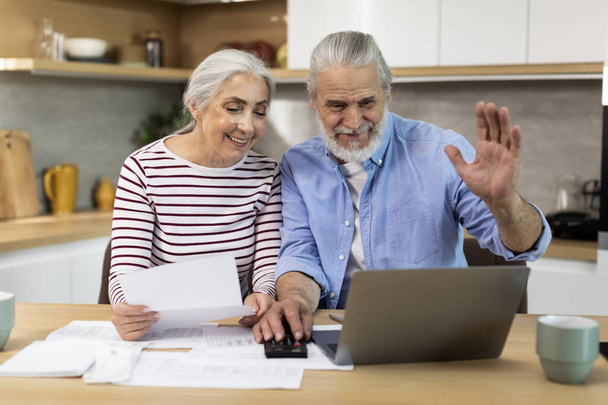 Happy Senior Spouses Making Video Call With Accountant While Using Laptop In Kitchen, Joyful Elderly Husband And Wife Waving At Camera And Checking Financial Papers, Planning Family Budget Together - Φωτογραφία, εικόνα