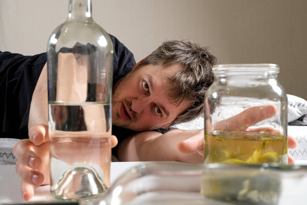Ein betrunkener Mann mit Alkoholsucht nimmt eine Flasche Wodka und Sole mit eingelegten Essiggurken - Foto, Bild