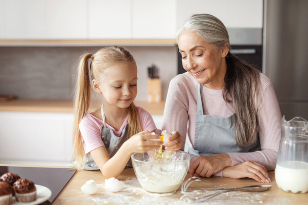 Glad smiling little girl and senior woman in aprons make dough, break egg in minimalist kitchen interior. Cooking lesson baking, household chores together, prepare food for family at home due covid-19 - 写真・画像
