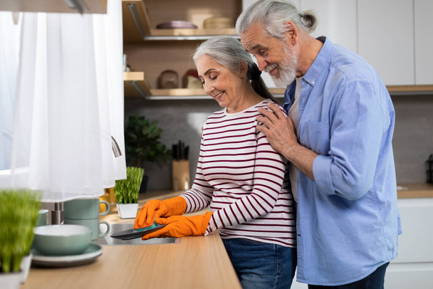 Portrait Of Happy Senior Spouses Washing Dishes In Kitchen After Lunch, Loving Elderly Husband Embracing Wife From The Back While She Doing Domestic Chores, Side View With Free Space - Zdjęcie, obraz