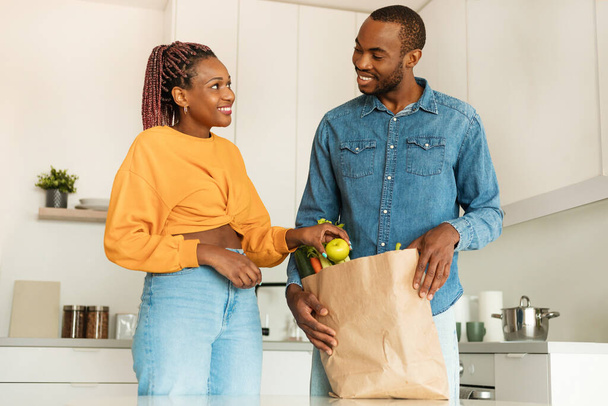 Ordering food online. Happy african american couple arriving from supermarket with grocery bag and unpacking in kitchen, looking at each other and smiling - Fotografie, Obrázek