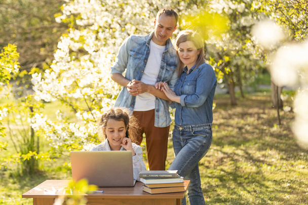 Happy family of four parents and cute little girl studying on laptop, enjoy using laptop watching cartoons, make internet video call or shopping online looking at computer screen sit together outdoor. - Photo, Image