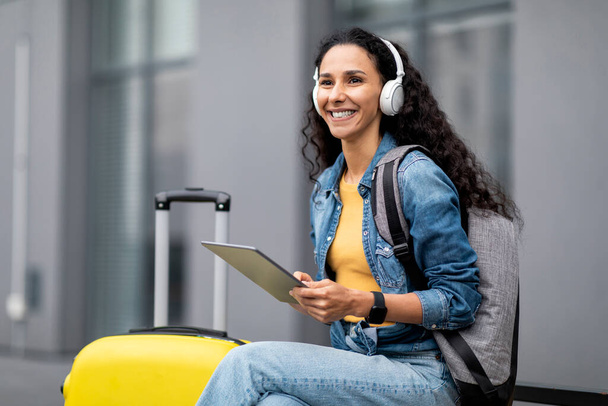 Happy young brunette lady with backpack and suitcase tourist using digital tablet and wireless headphones, sitting on bench outdoors, looking at copy space and smiling. Entertainment while travelling - Foto, afbeelding