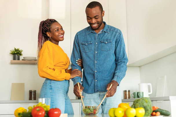 Loving black man cooking healthy dinner for his beautiful wife, preparing fresh salad in white cozy kitchen interior, copy space. Love, affectionate, family lifestyle concept - Foto, Bild