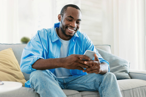 Excited black man chatting on smartphone, watching video, playing game while resting on sofa in living room interior. Free time for blog at home, new app, advertisement and offer - Photo, image