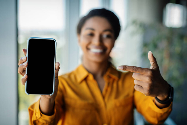 Great offer. Black businesswoman holding and pointing finger at cellphone with empty screen, promoting app or website, advertising product or service, standing in office - Foto, Imagen