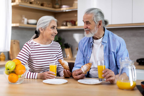 Портрет веселої літньої пари, що сніданок разом на кухні Інтер'єр, щасливі старші подружжя їдять круасани і п'ють апельсиновий сік, насолоджуючись проводити час вдома один з одним
 - Фото, зображення
