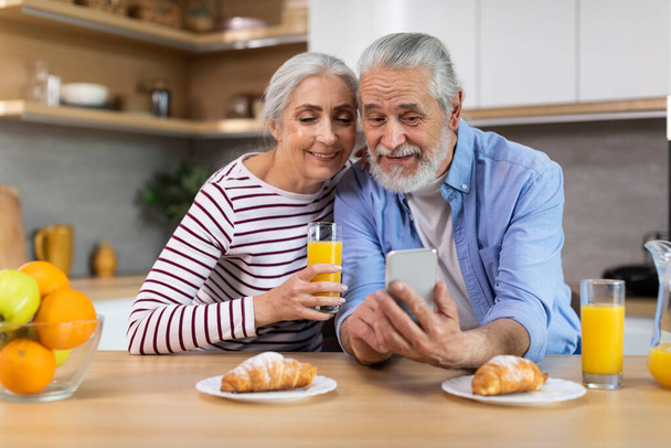Portrait Of Happy Senior Spouses Using Smartphone During Breakfast In Kitchen, Married Elderly Couple Browsing Internet On Mobile Phone Or Checking New App, Closeup Shot With Free Space - Foto, imagen