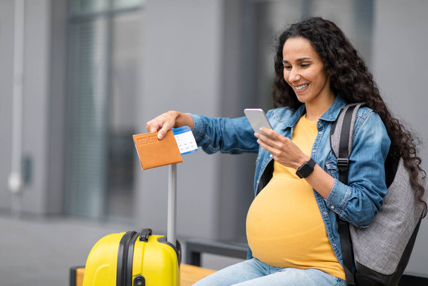 Giving birth in foreign country concept. Happy young long-haired brunette pregnant woman travelling alone, sitting on bench with backpack next to baggage, using cell phone and smiling, copy space - Photo, Image