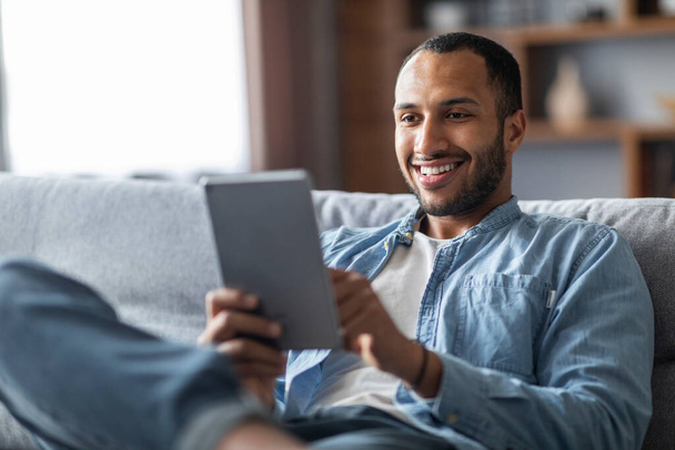 Handsome smiling black male relaxing with digital tablet at home, young african american man resting on couch and using modern gadget, browsing internet or shopping online, closeup shot - 写真・画像