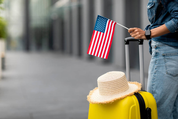 Unrecognizable woman with straw hat on yellow suitcase holding flag of the US, posing in airport, panorama with copy space. Travelling, tourism, vacation, holidays, education abroad concept - Фото, изображение
