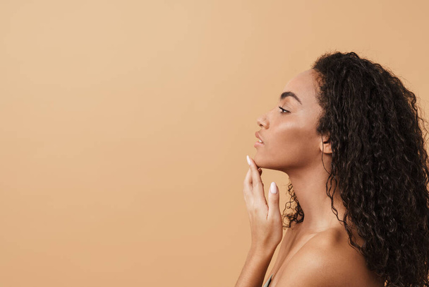 Shirtless black woman with wavy hair touching her facial skin isolated over beige background - Photo, Image