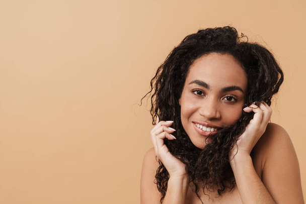 Shirtless black woman smiling and holding her hair isolated over beige background - Фото, изображение