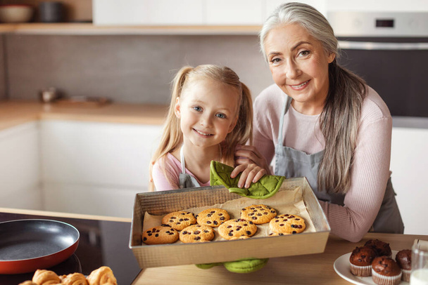 Smiling little girl and senior female in aprons show homemade fresh cookies in kitchen interior. Household chores together, prepare baking and cooking food for family at home. Sweets ready to eat - Foto, Imagen