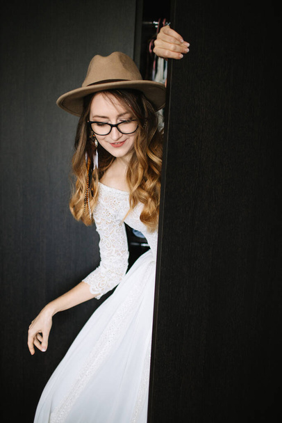 The bride in a white dress with lace sleeves, a brown hat, with feathers in her hair looks out from behind a black door and smiles. Graceful hand curve. Bride on a black background. - Fotoğraf, Görsel
