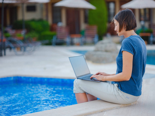 Young business woman working at computer by hotel pool. Young lady downshifter working at laptop and enjoys and relaxed environment, working day. Online freelance work on vacation. - Photo, image