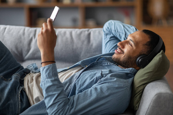 Favorite Leisure. Happy Black Guy In Wireless Headphones Listening Music On Smartphone While Relaxing On Couch At Home, Smiling African American Man Resting On Sofa And Enjoying Favorite Playlist - Photo, image