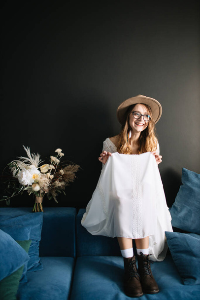 The bride in a white dress with lace, black boots, a brown hat and glasses sits on the back of a blue sofa, holds the hem of her dress and laughs. Near the wedding bouquet in boho style. - Fotó, kép