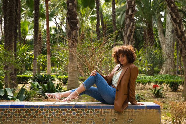 Hermosa mujer con el pelo rizado sentado en un banco en un parque con grandes árboles en una ciudad europea. La mujer está vestida con ropa moderna y de moda. Concepto de viaje y vacaciones, descanso y relajación. - Foto, imagen