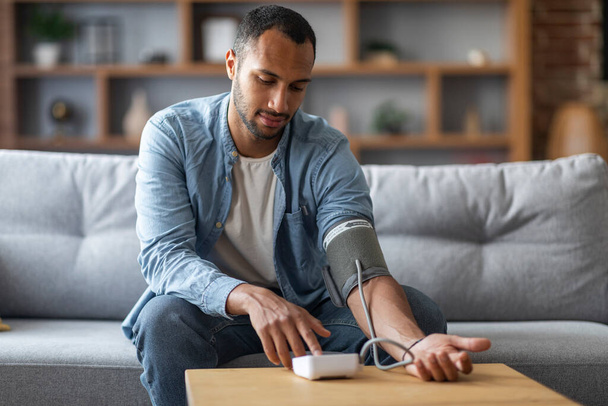 Young Black Man Sitting On Couch And Checking Blood Pressure With Upper Arm Monitor, Millennial African American Guy Measuring Arterial Tension While Relaxing In Living Room At Home, Copy Space - Photo, image