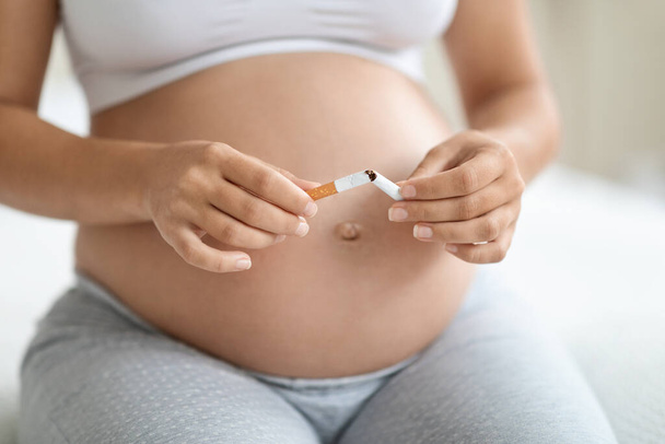 Healthy life choice during pregnancy, stop smoking concept. Unrecognizable pregnant woman sitting on bed at home, breaking cigarette on halves, cropped photo, free space - Fotó, kép