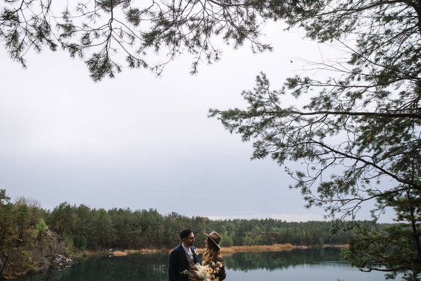 The bride and groom are standing near the lake in the forest, looking at each other and smiling. The bride holds a wedding bouquet in boho style. Wedding in the forest. - Foto, immagini