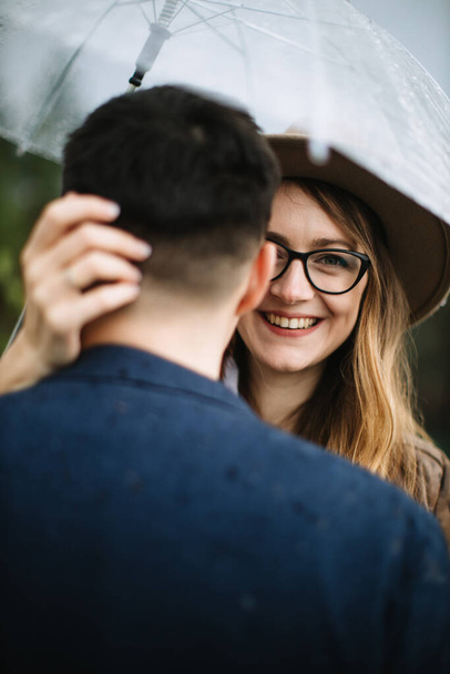 Lovers stand under an umbrella in the rain against each other. The girl's hand lies on the back of the guy's head, the girl in glasses and a hat laughs. - Φωτογραφία, εικόνα