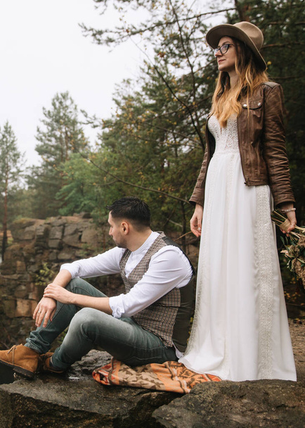 The bride and groom are on a rock with a pine forest. The groom sits on a Navajo rug on a rock. The bride in a white dress, leather jacket, and hat holds a wedding bouquet. Wedding in nature. - Foto, Imagen