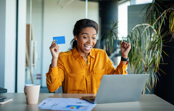 Overjoyed black female office worker holding credit card in front of laptop and making YES gesture, shopping while sitting at workplace. Cool online promo concept - Fotoğraf, Görsel